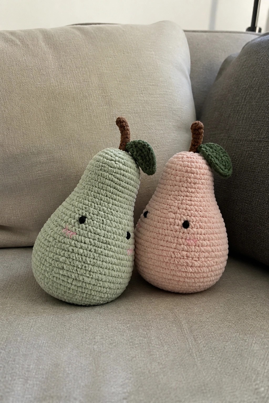 Two crocheted pears, mint green and blush pink, with cute faces on a couch.