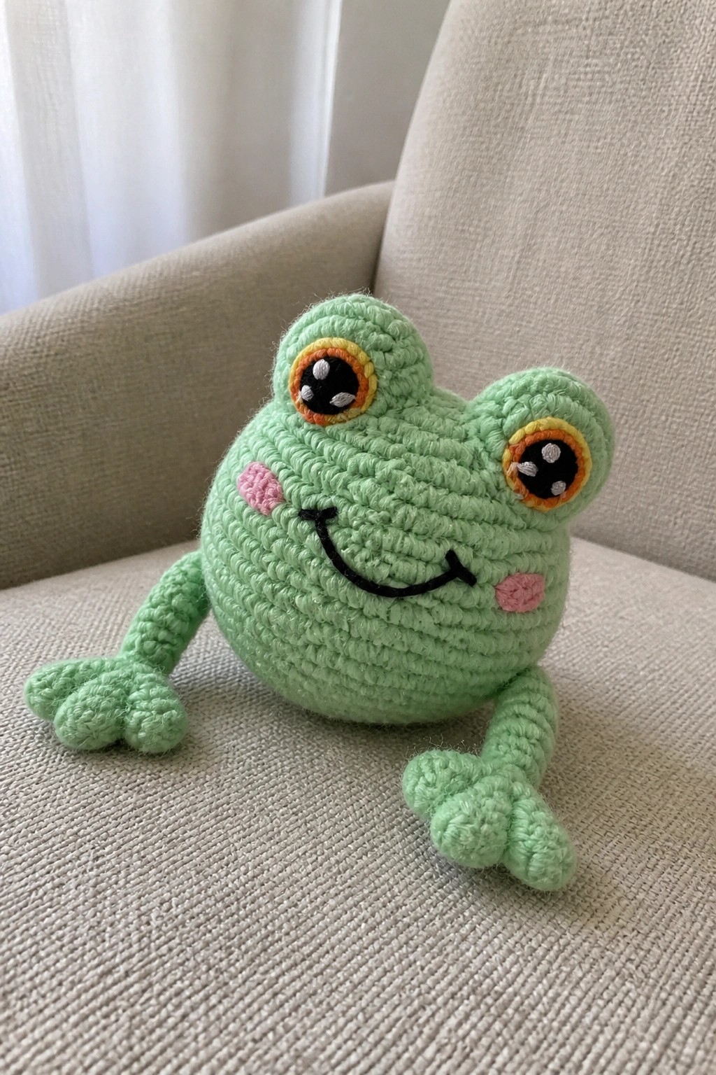 A light green crocheted frog amigurumi with large eyes, pink cheeks, and a black smile sits on a beige fabric chair.