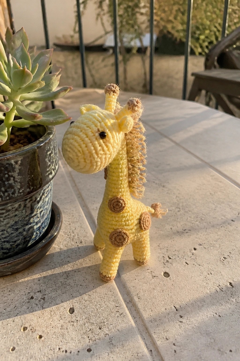 Crocheted yellow giraffe toy with brown spots beside potted succulent on sunny table.