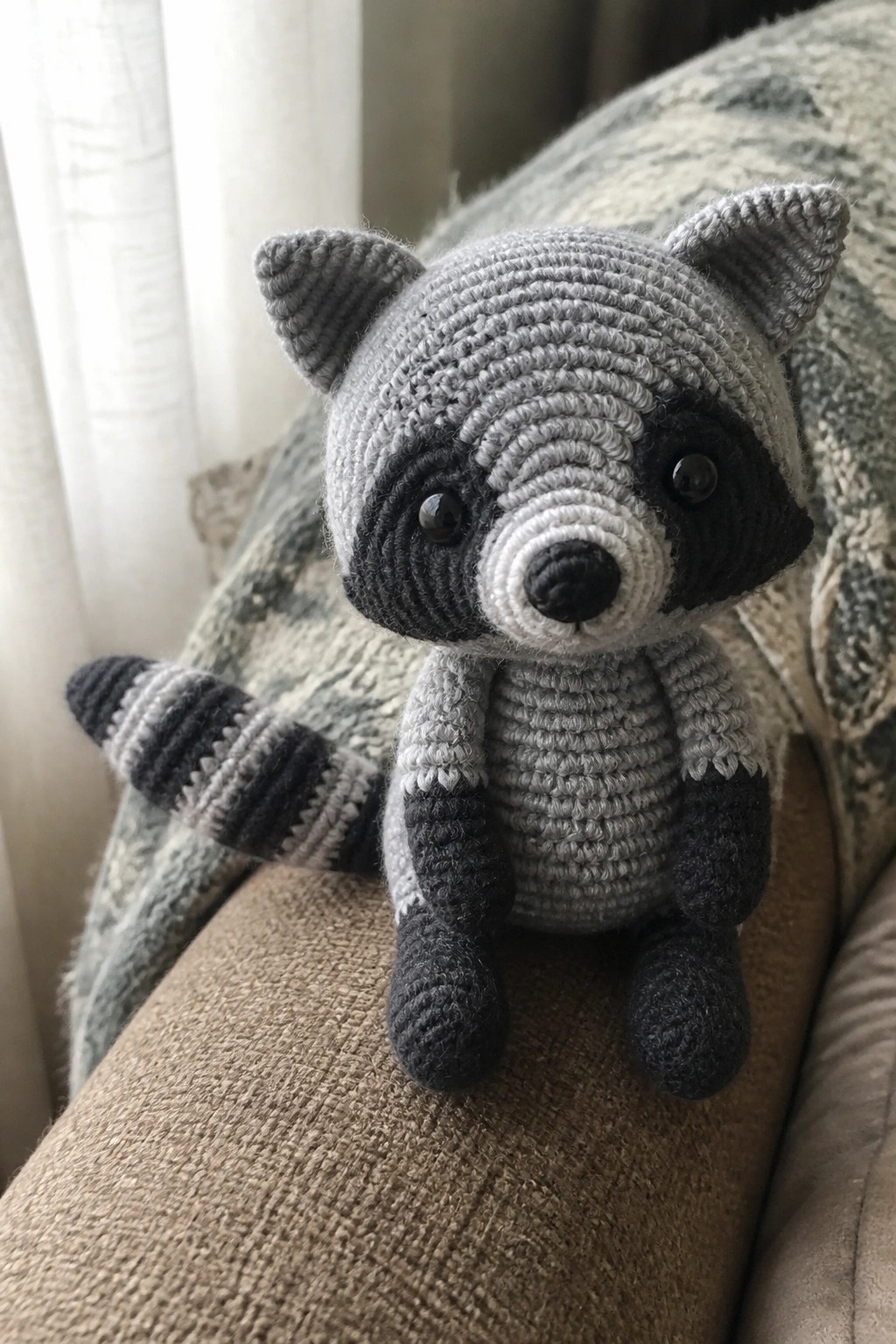 Crocheted gray raccoon toy with black mask and striped tail on couch