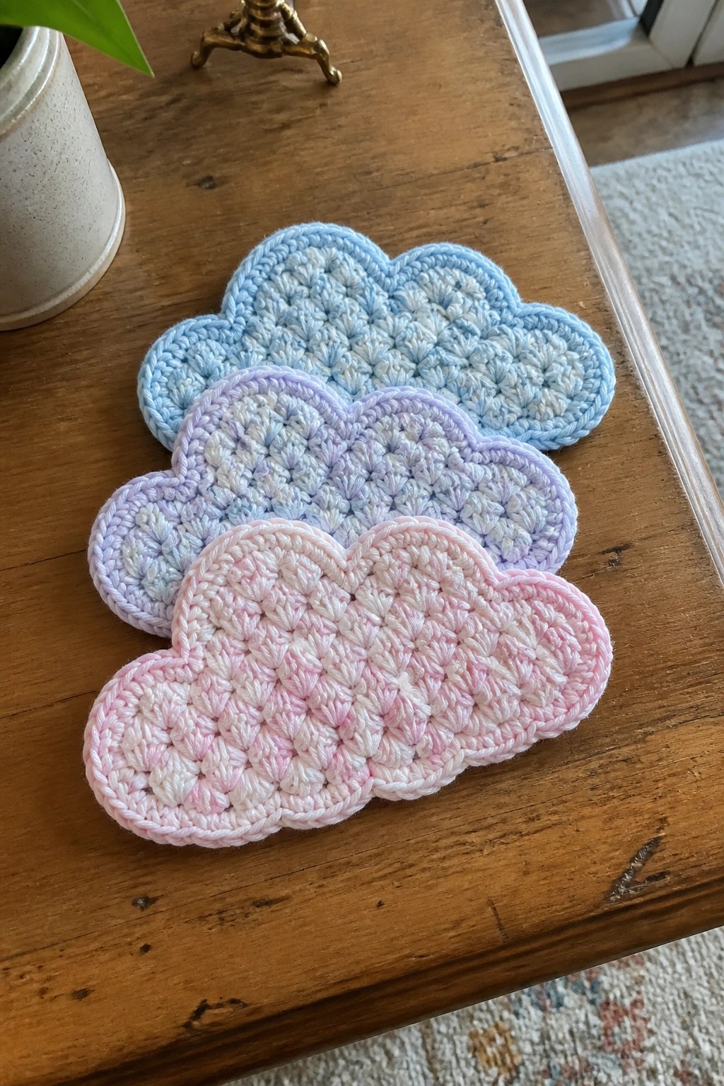 Three pastel crocheted cloud coasters in blue, purple, and pink on wood table