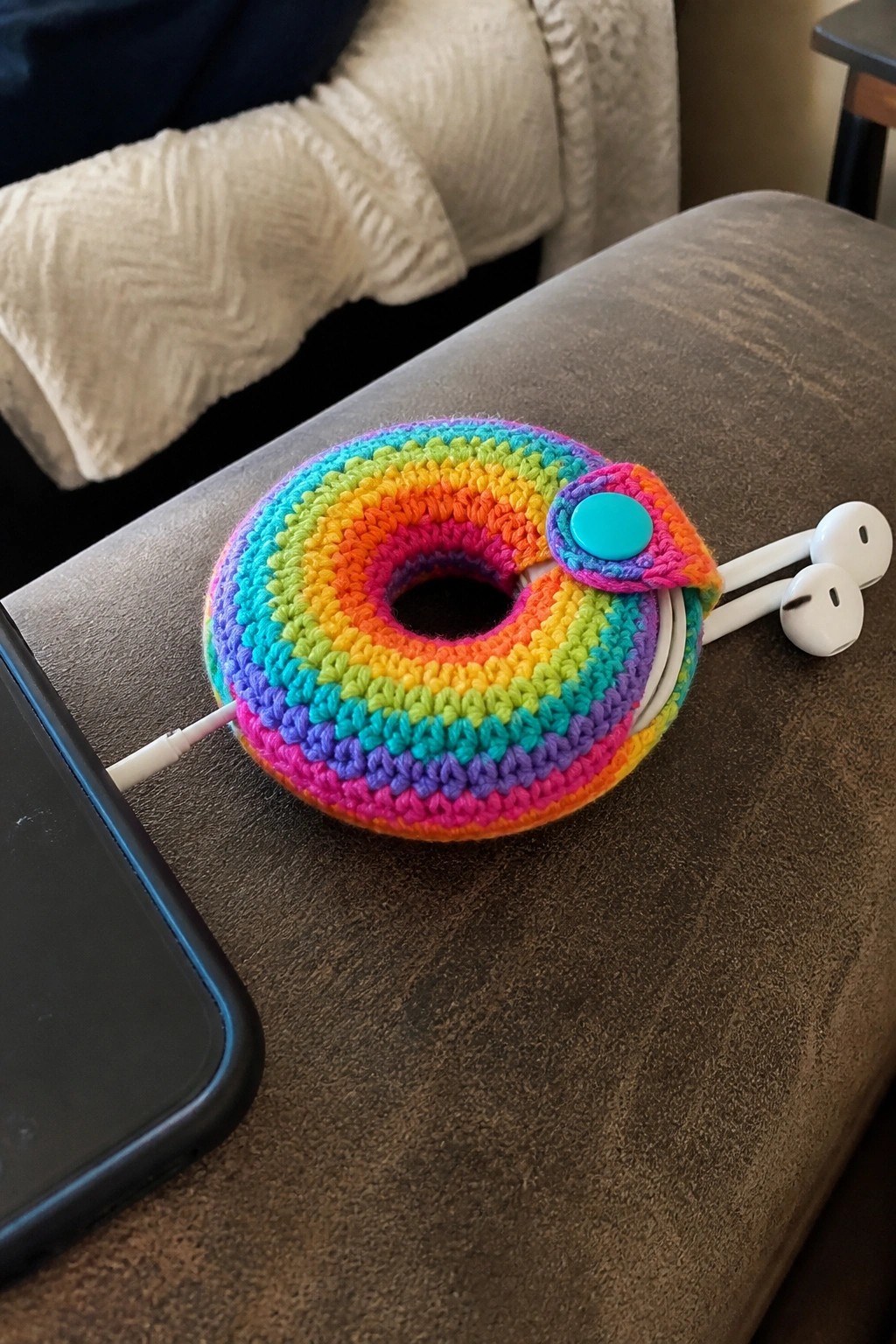 Rainbow crocheted donut pouch with button holding white earbuds plugged into phone.