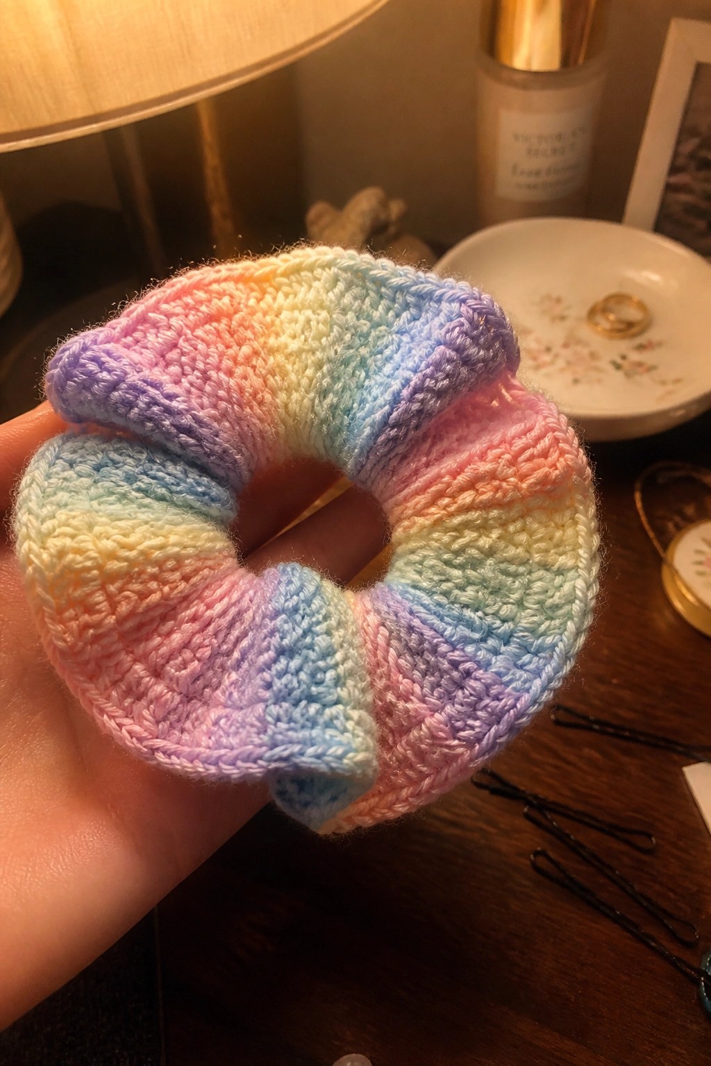 Hand holding a rainbow crocheted scrunchie on a wooden surface with lamp nearby