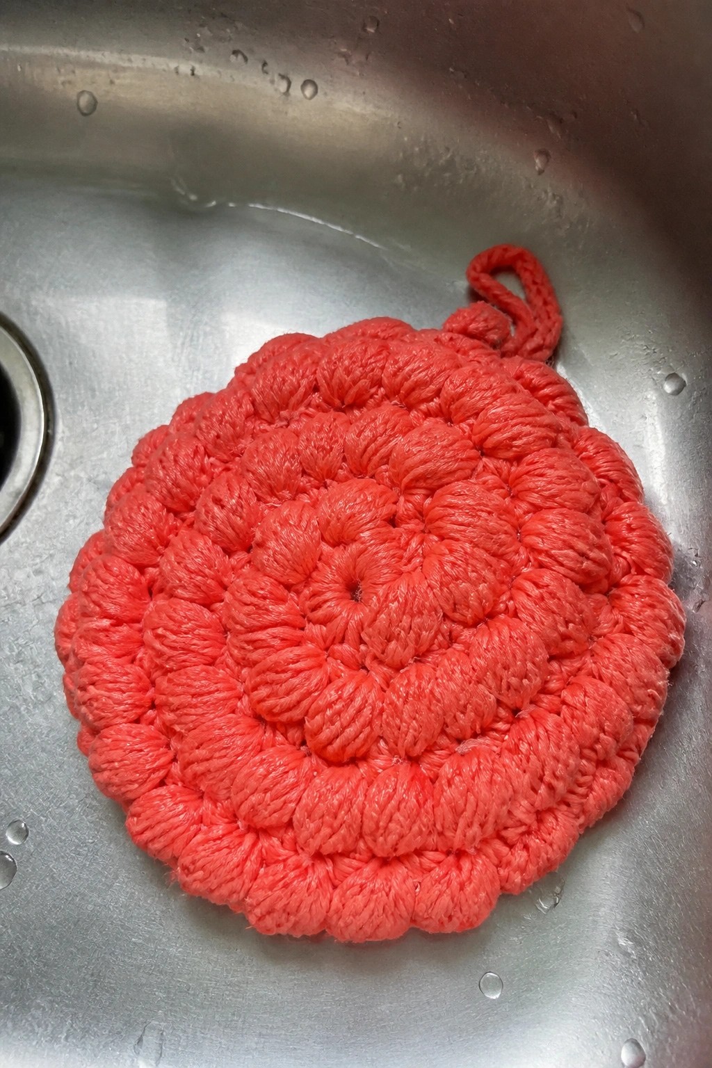 Red crocheted circular scrubber with loop in wet stainless steel sink