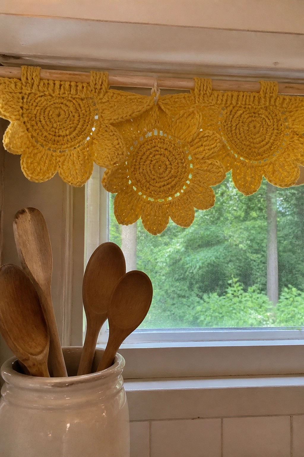 Yellow crocheted sunflower valance hangs over window above jar of wooden spoons