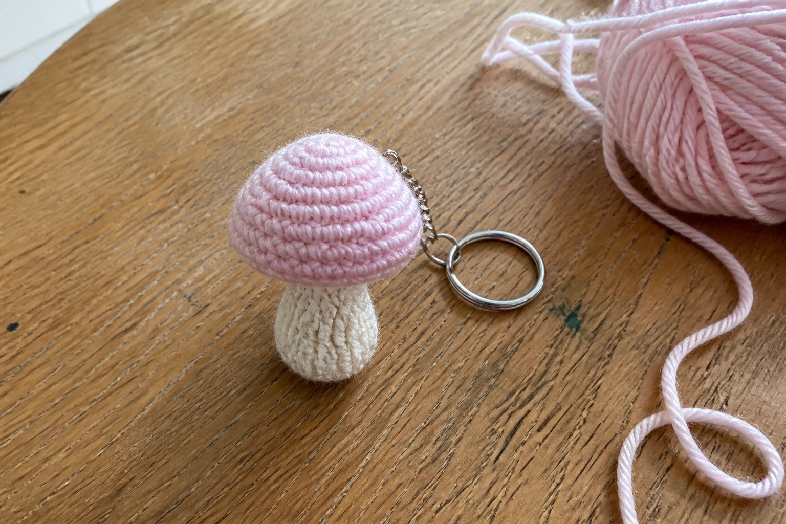 Pink crocheted mushroom keychain on wooden table beside pink yarn ball