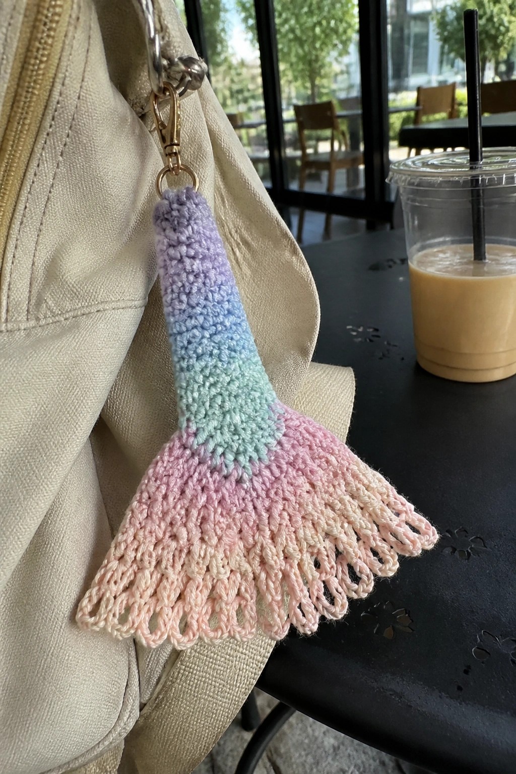 Pastel crocheted mermaid tail keychain on beige bag beside iced coffee cup
