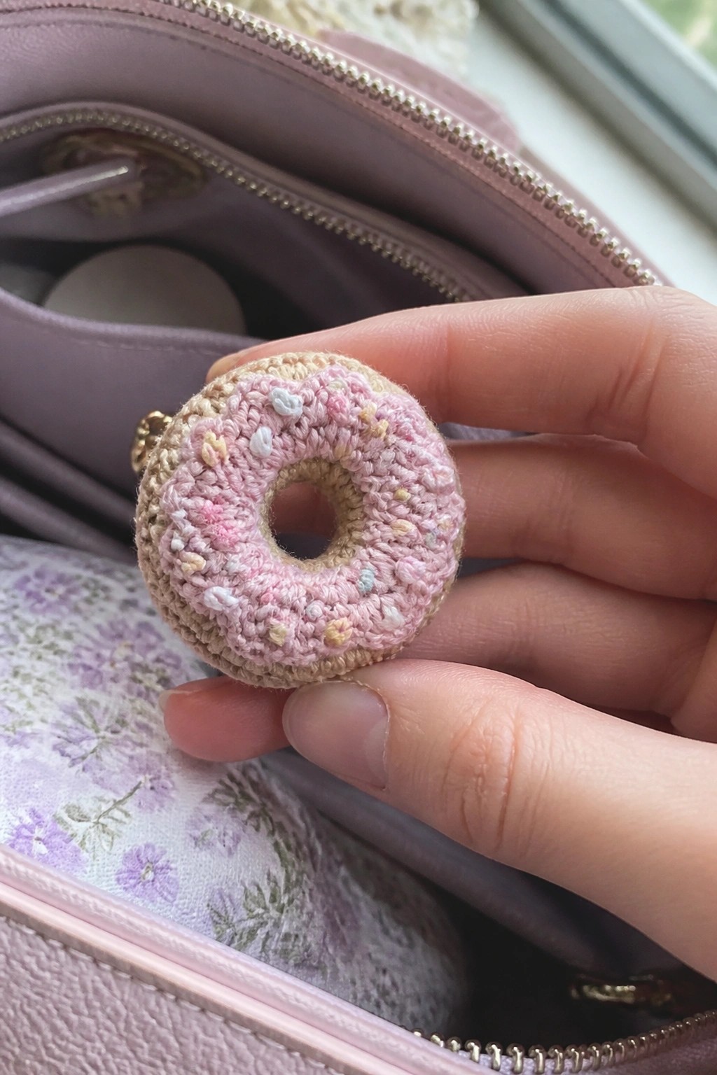 Hand holding pink crocheted donut with sprinkles over open floral-lined pink bag