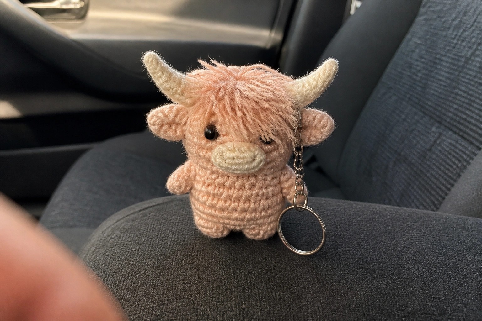 Hand holding pink crocheted Highland cow keychain with horns on car seat