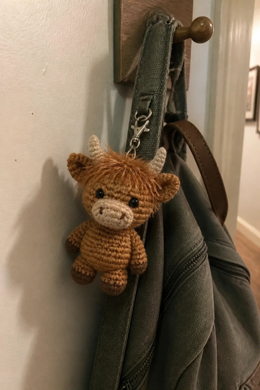 Crocheted brown cow keychain clipped to green backpack on wooden wall hook.