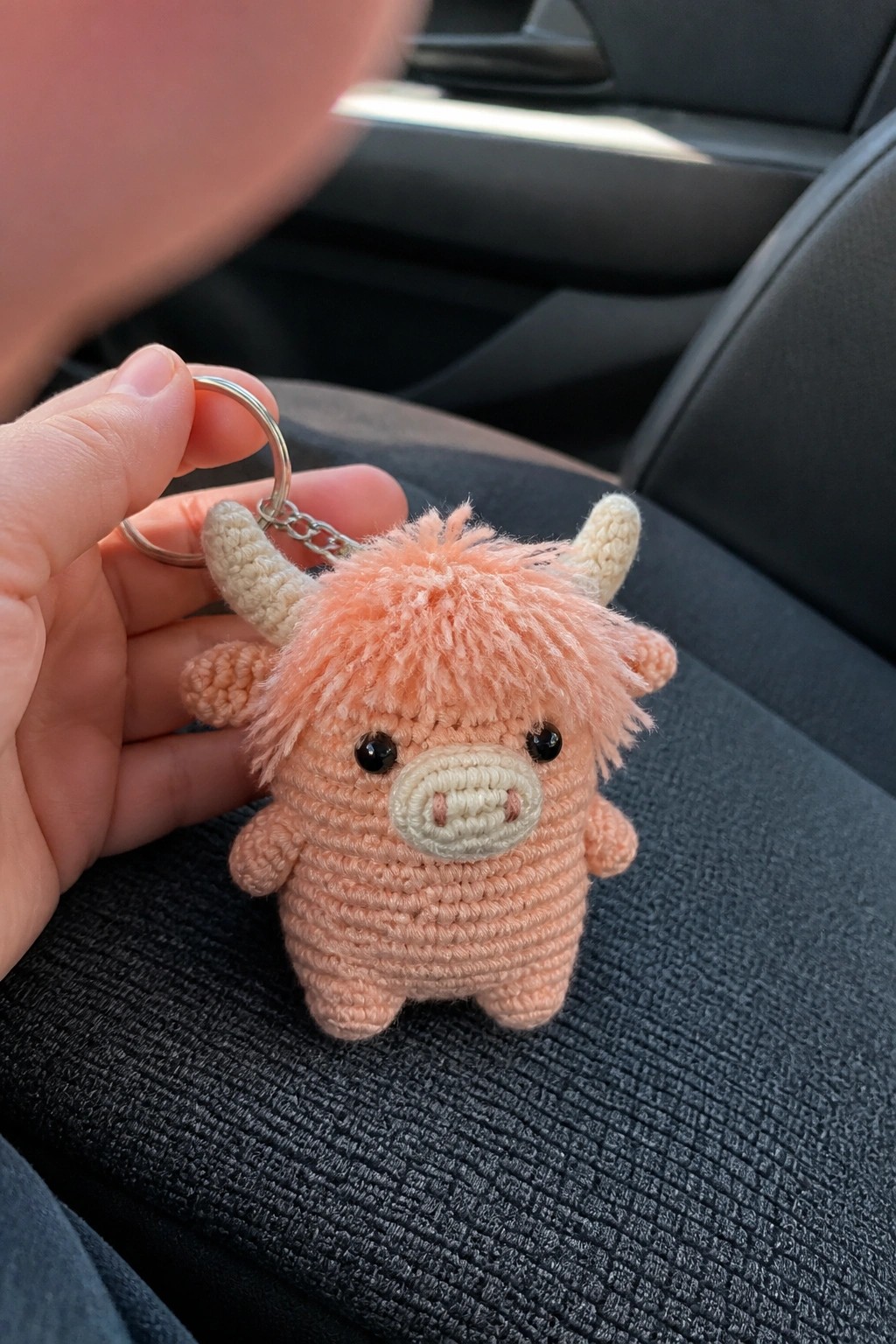 Hand holding pink crocheted Highland cow keychain with horns on car seat