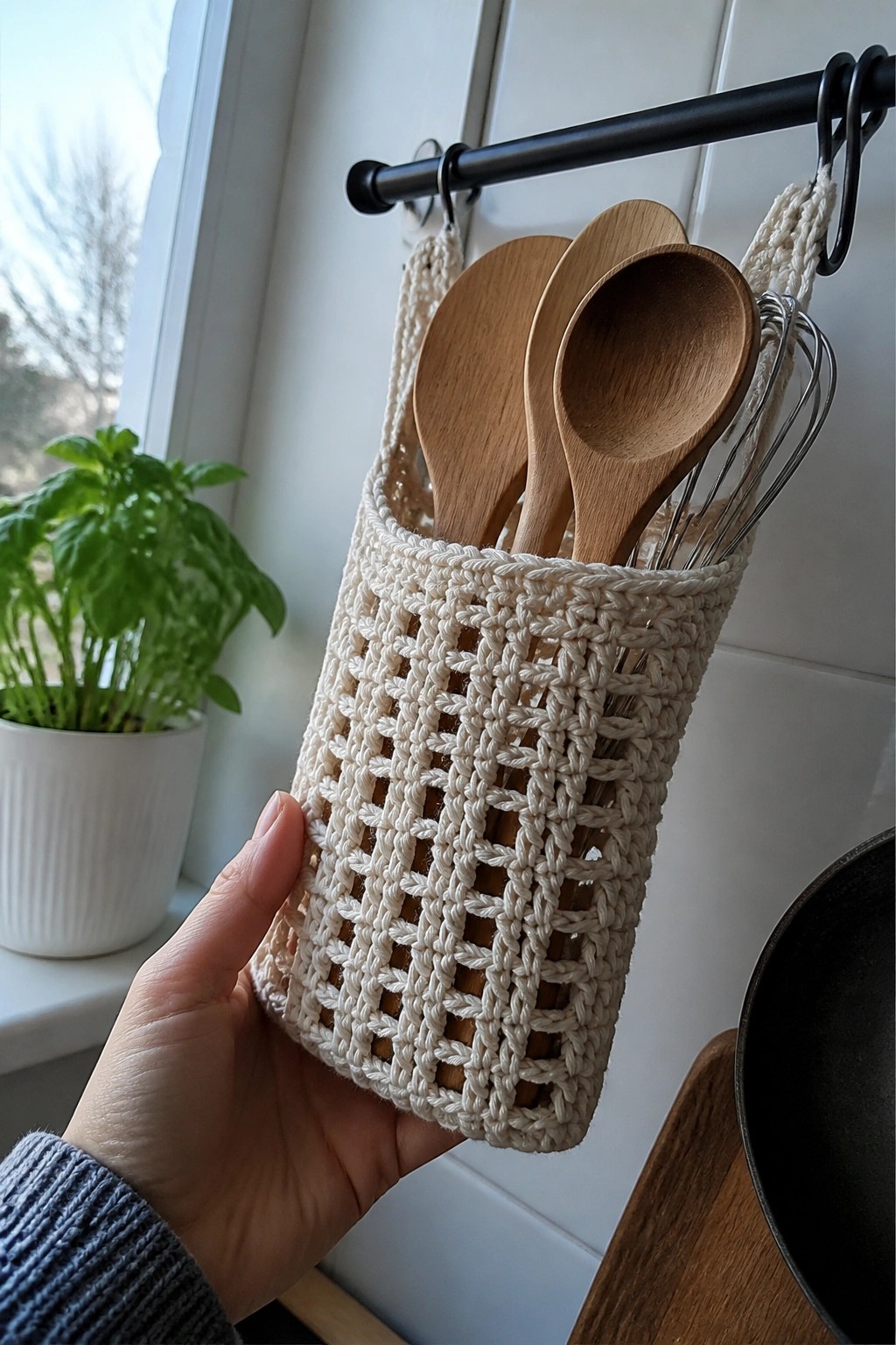 Hand holding off-white crocheted basket with wooden spoons and whisk on rod