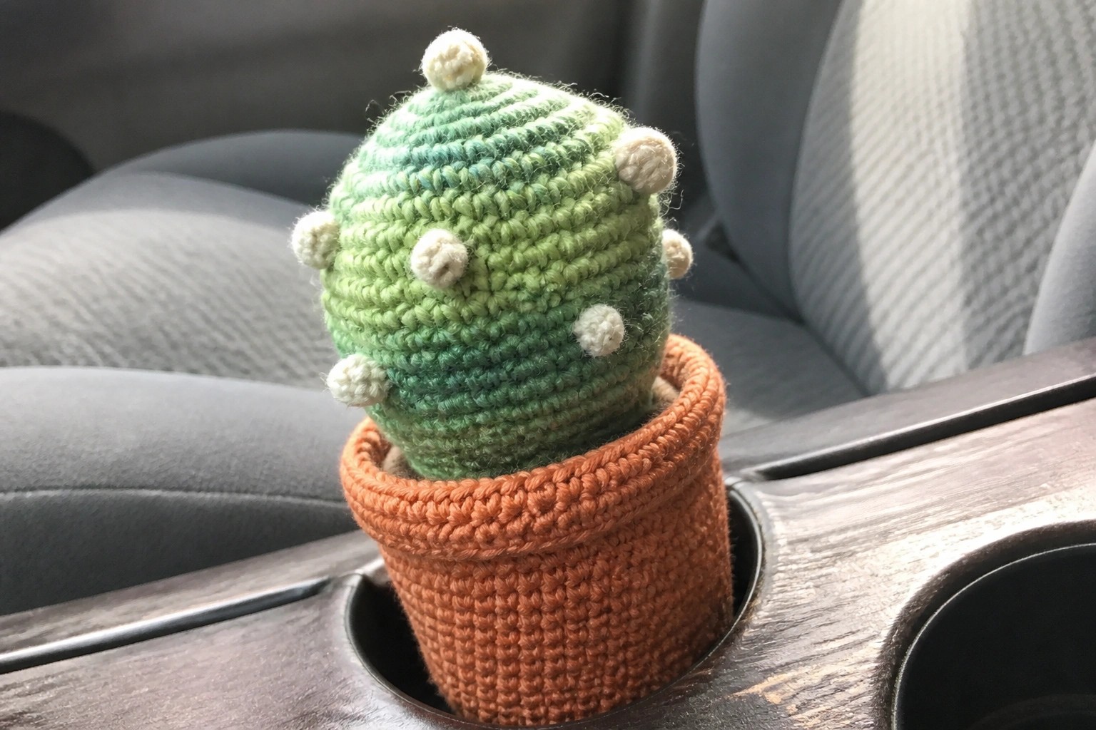 A small crocheted green cactus with light spots sits in an orange crocheted pot inside a car cup holder.