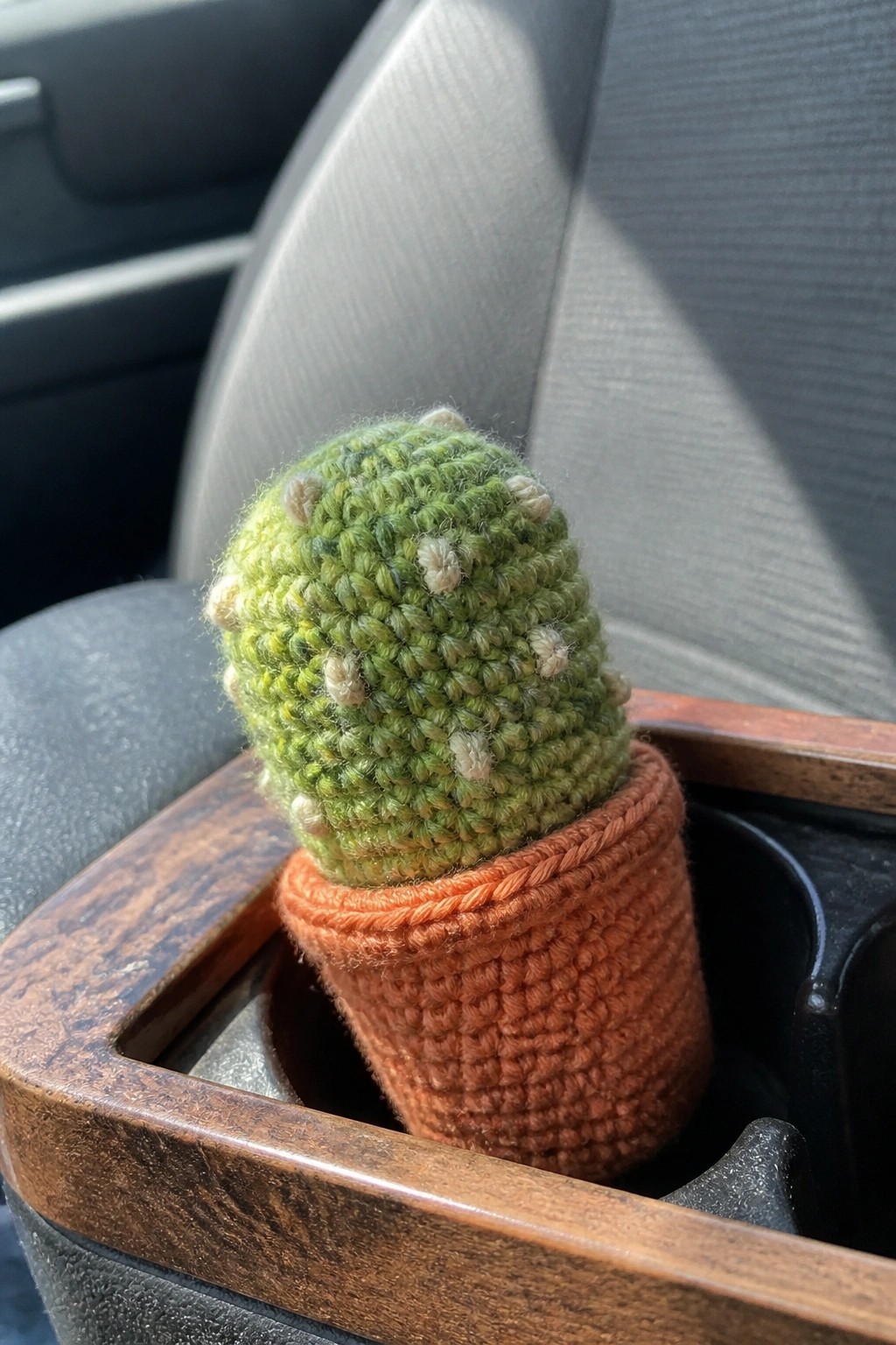 A small crocheted green cactus with light spots sits in an orange crocheted pot inside a car cup holder.
