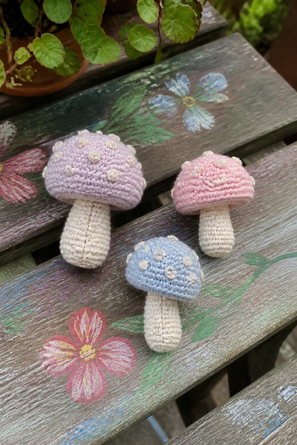 Three pastel crocheted mushrooms on a flower-painted wooden surface.