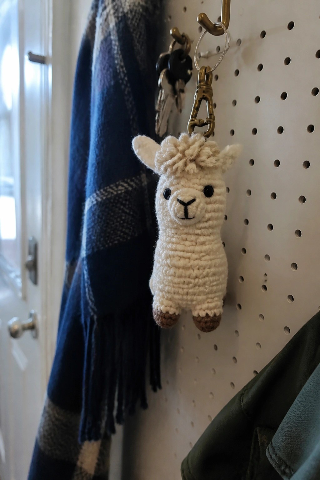Crocheted white llama keychain with brown feet hangs on pegboard beside plaid scarf.