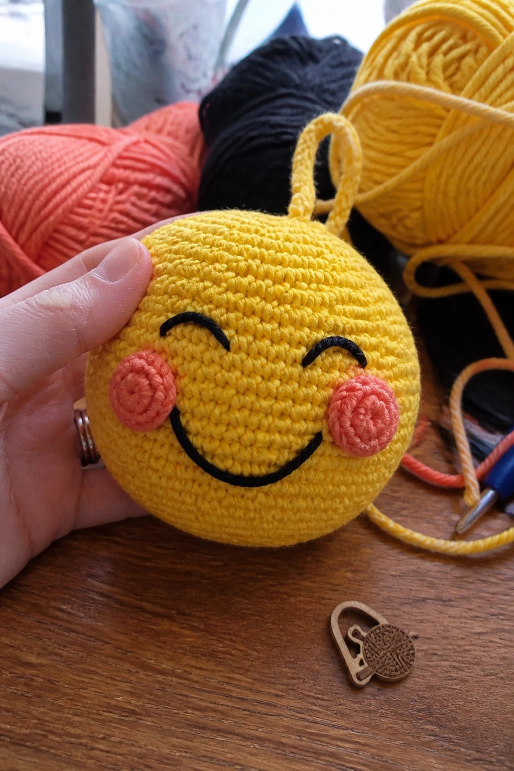 A hand holds a yellow crocheted smiley face ball with black embroidered eyes and mouth, pink cheeks, and two yellow yarn loops on top, with yarn balls and a crochet hook visible on a wooden table.