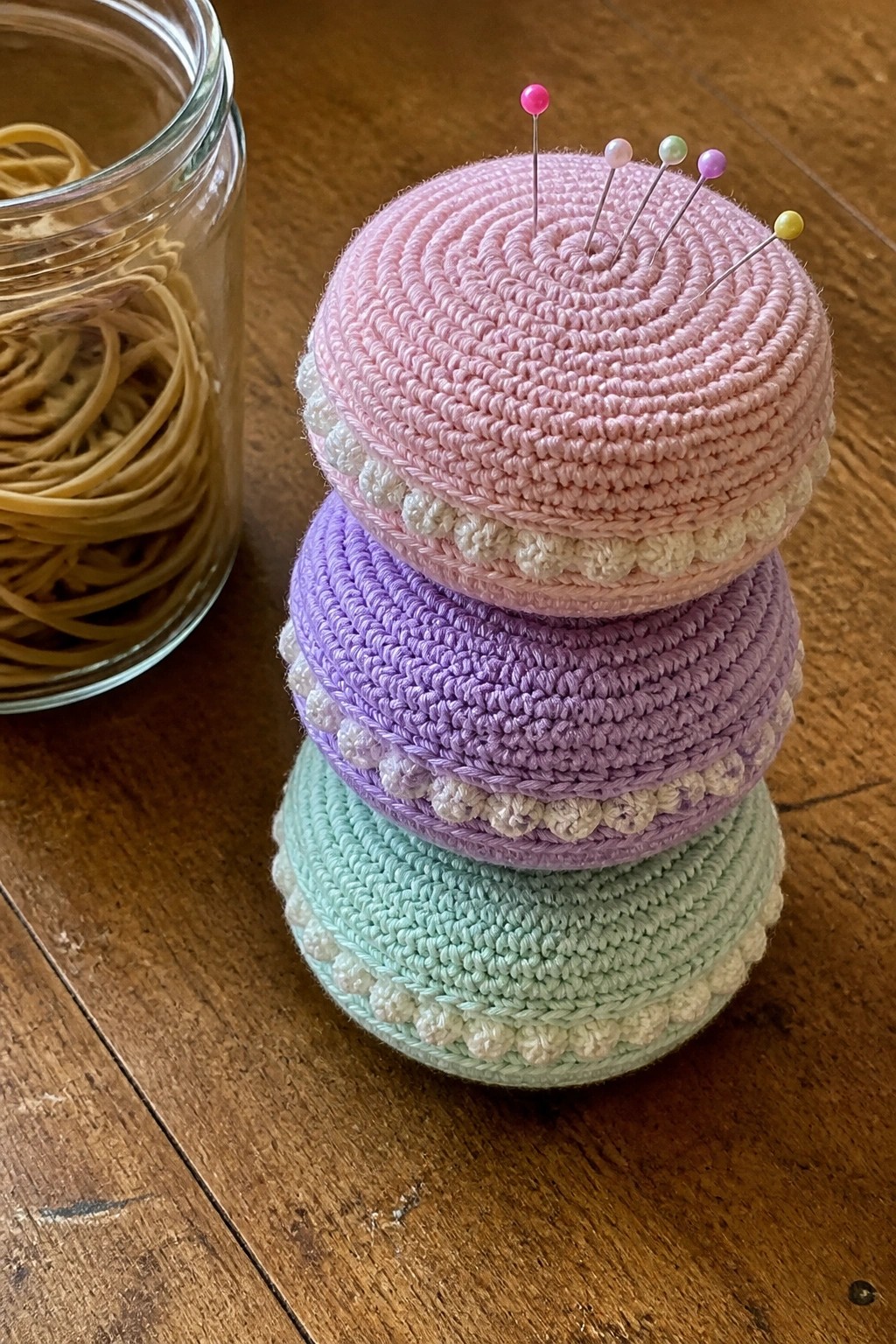 Stacked pastel crocheted pincushions with pins beside rubber band jar on wood
