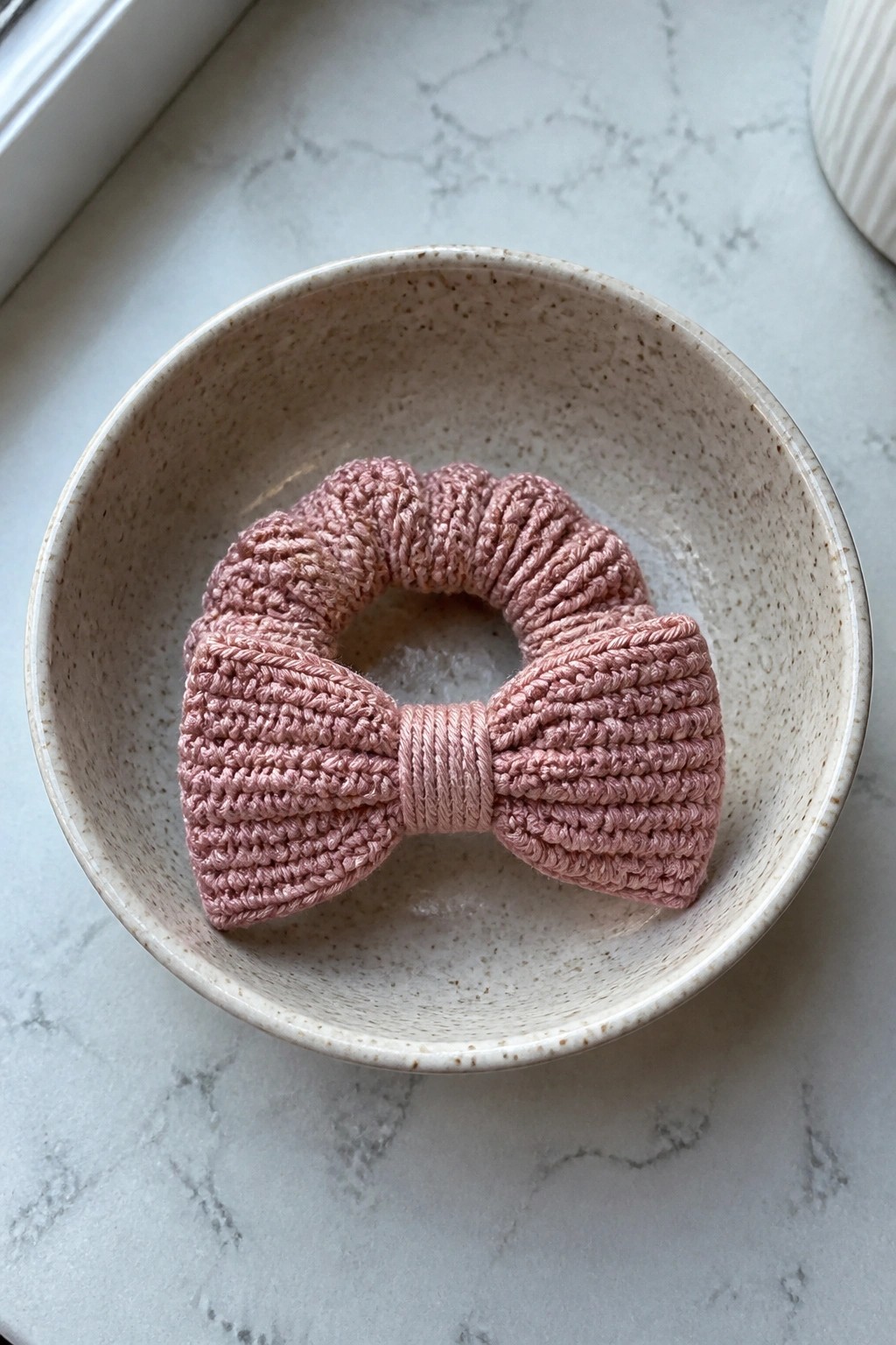 Pink crocheted bow scrunchie in a speckled beige ceramic bowl on marble