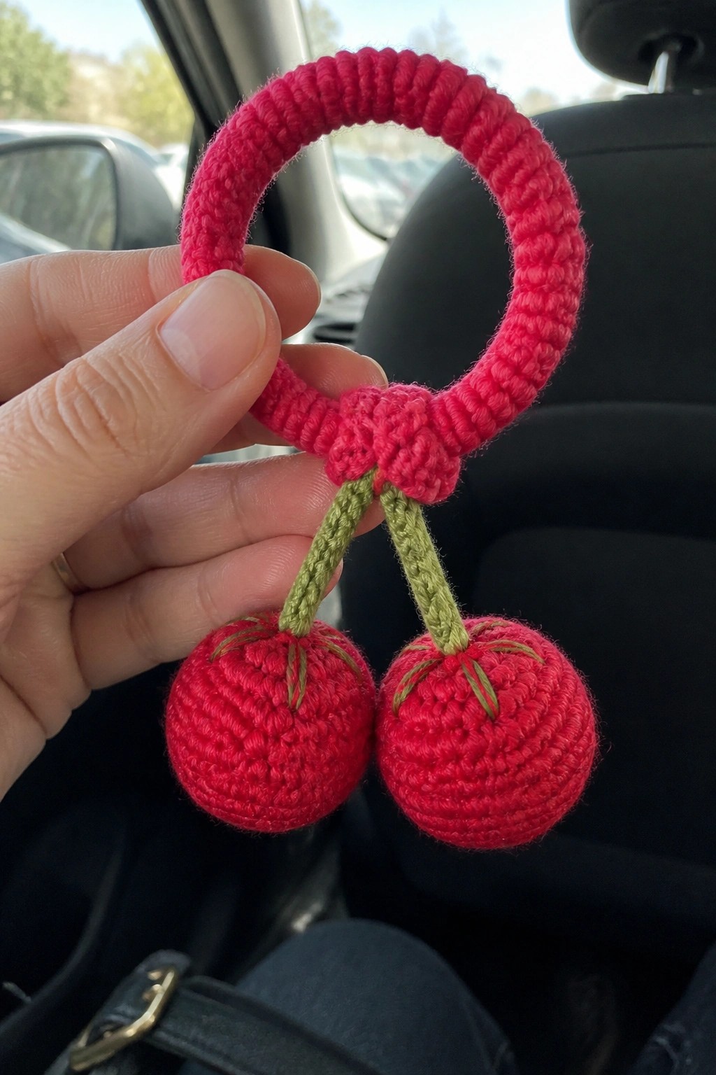 Hand holding crocheted pink ring with two red cherry ornaments in car.