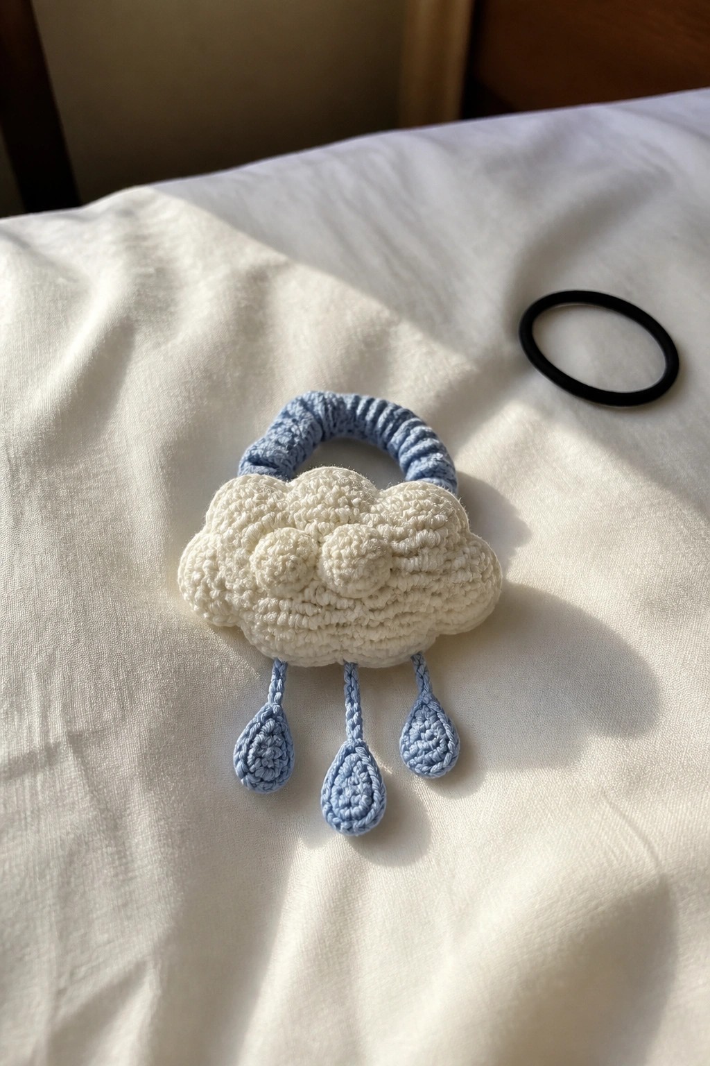 White crocheted cloud with three blue raindrops on blue scrunchie atop white fabric