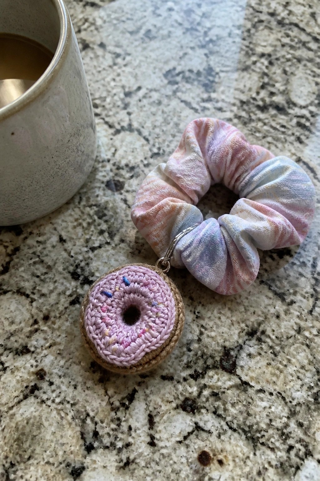 Pastel tie-dye scrunchie with crocheted pink donut charm beside coffee mug on marble.