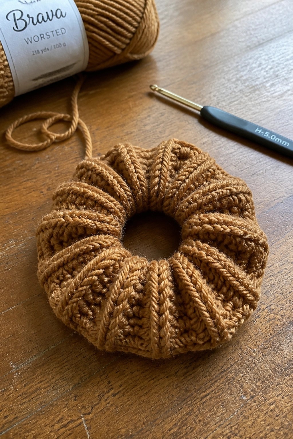 Brown crocheted scrunchie beside worsted yarn skein and 5mm hook on wood
