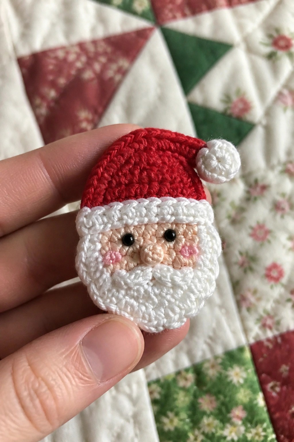 Hand holding miniature crocheted Santa face with red hat, white beard on quilt