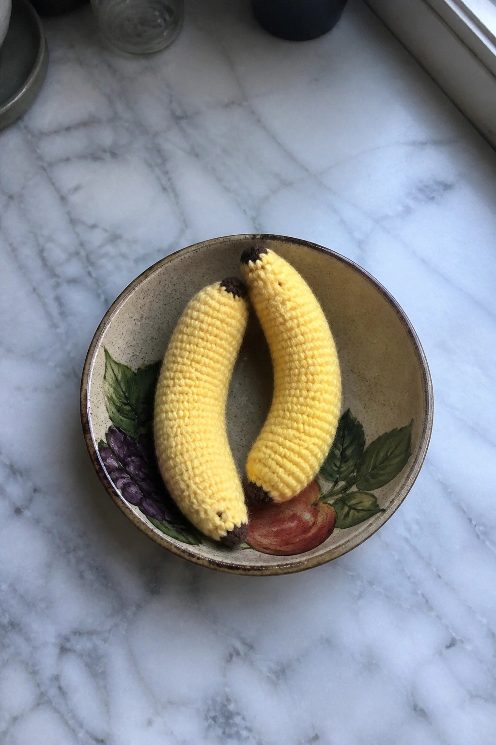 Two yellow crocheted bananas in a fruit-patterned ceramic bowl on marble countertop.