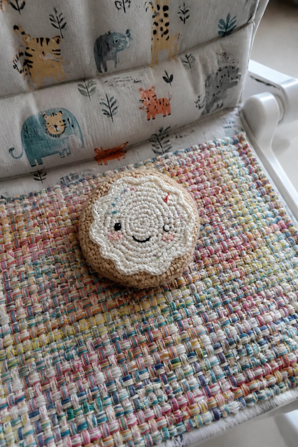 Crocheted sheep face on beige base atop colorful woven mat with animal prints behind.