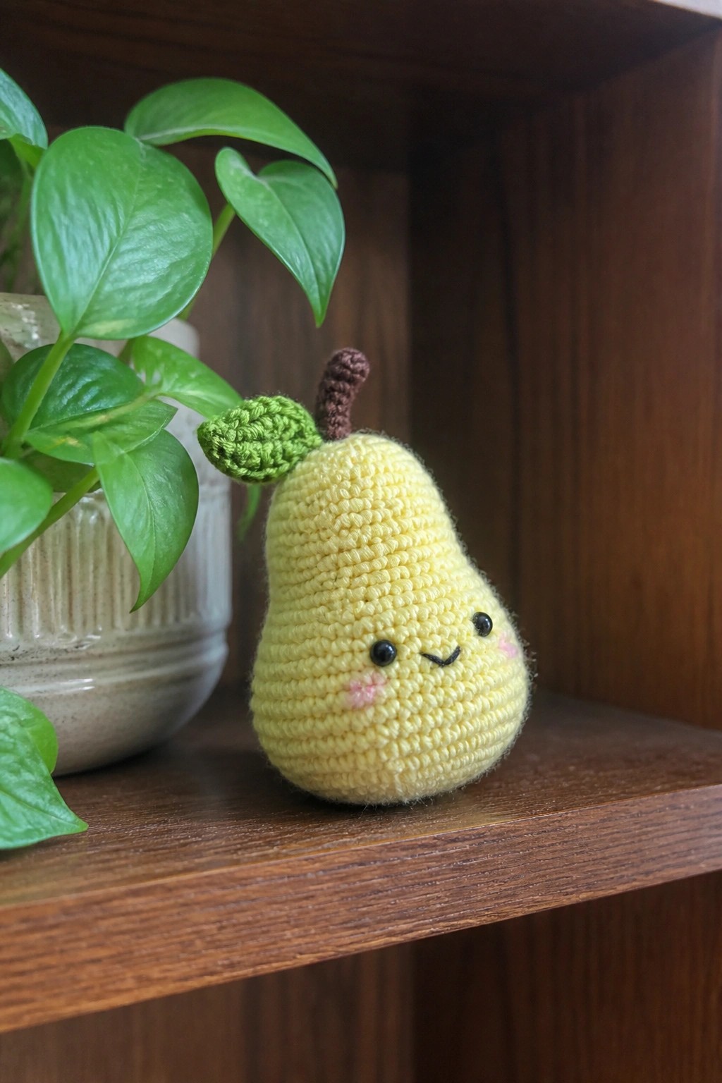 Yellow crocheted pear with smiling face on wooden shelf beside plant.