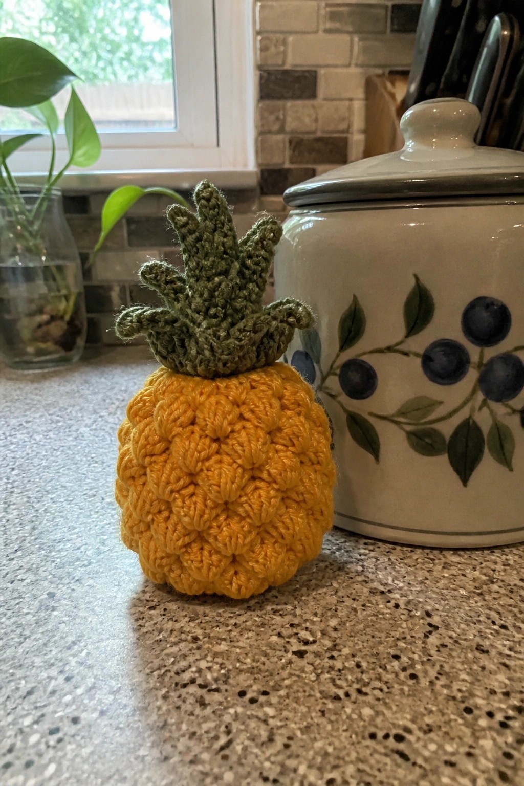 Crocheted yellow pineapple with green top on kitchen counter beside blueberry jar.