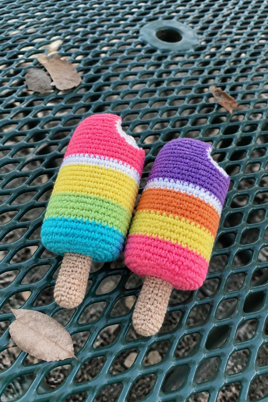 Two colorful crocheted rainbow popsicles on a green metal picnic table.