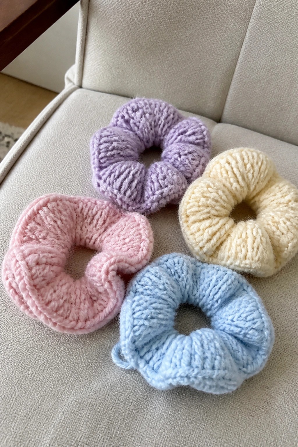 Four pastel knitted scrunchies in pink, purple, blue, and cream on beige fabric.