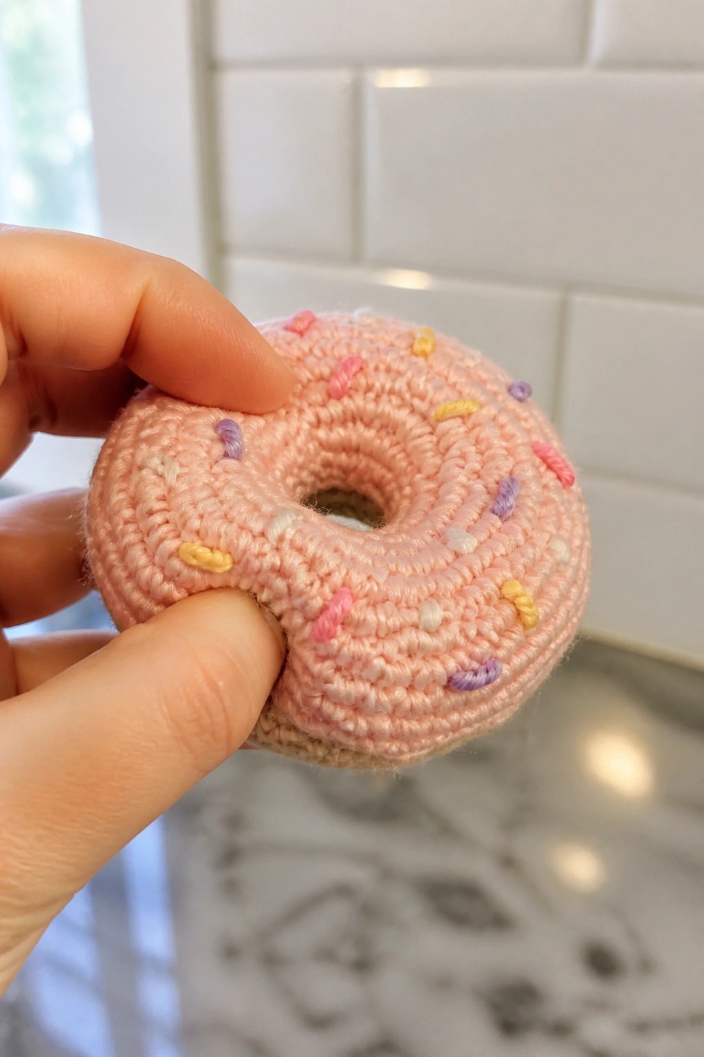 Hand holding pink crocheted donut with colorful yarn sprinkles on marble counter.
