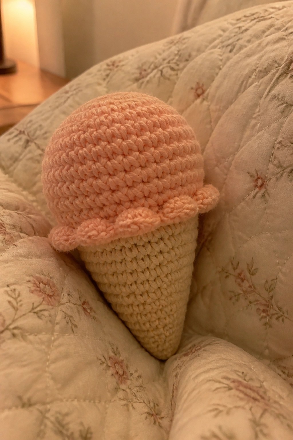 A crocheted pink ice cream scoop sits atop a beige cone on a floral quilt.