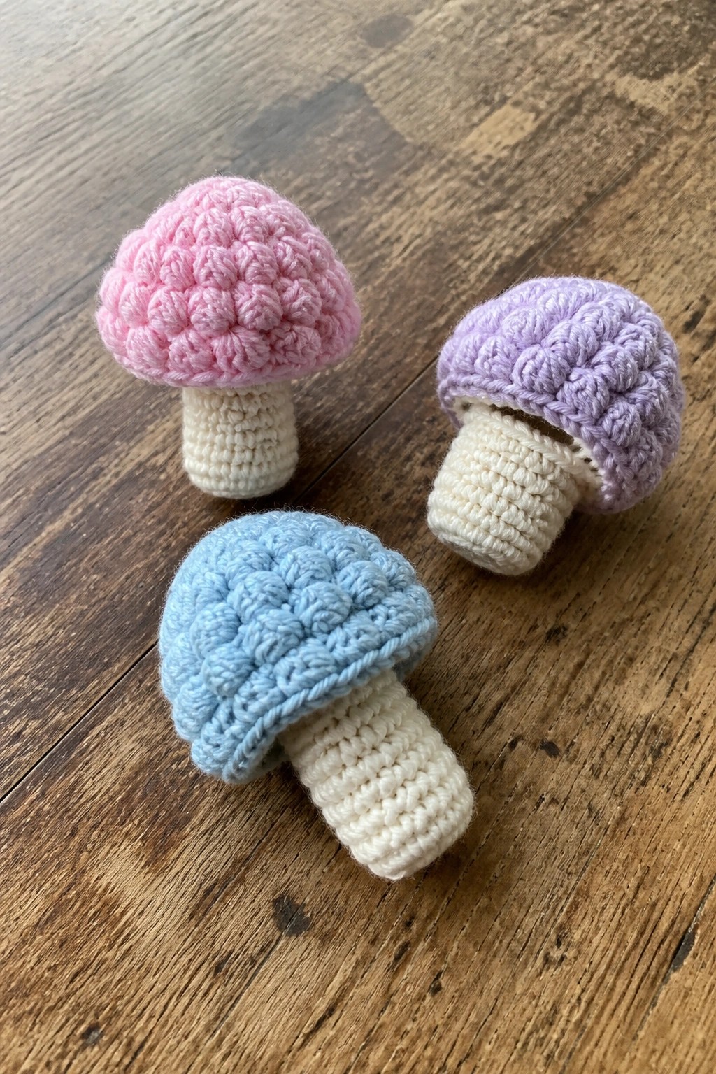 Three crocheted mushrooms with pink, purple, and blue caps on wooden surface.