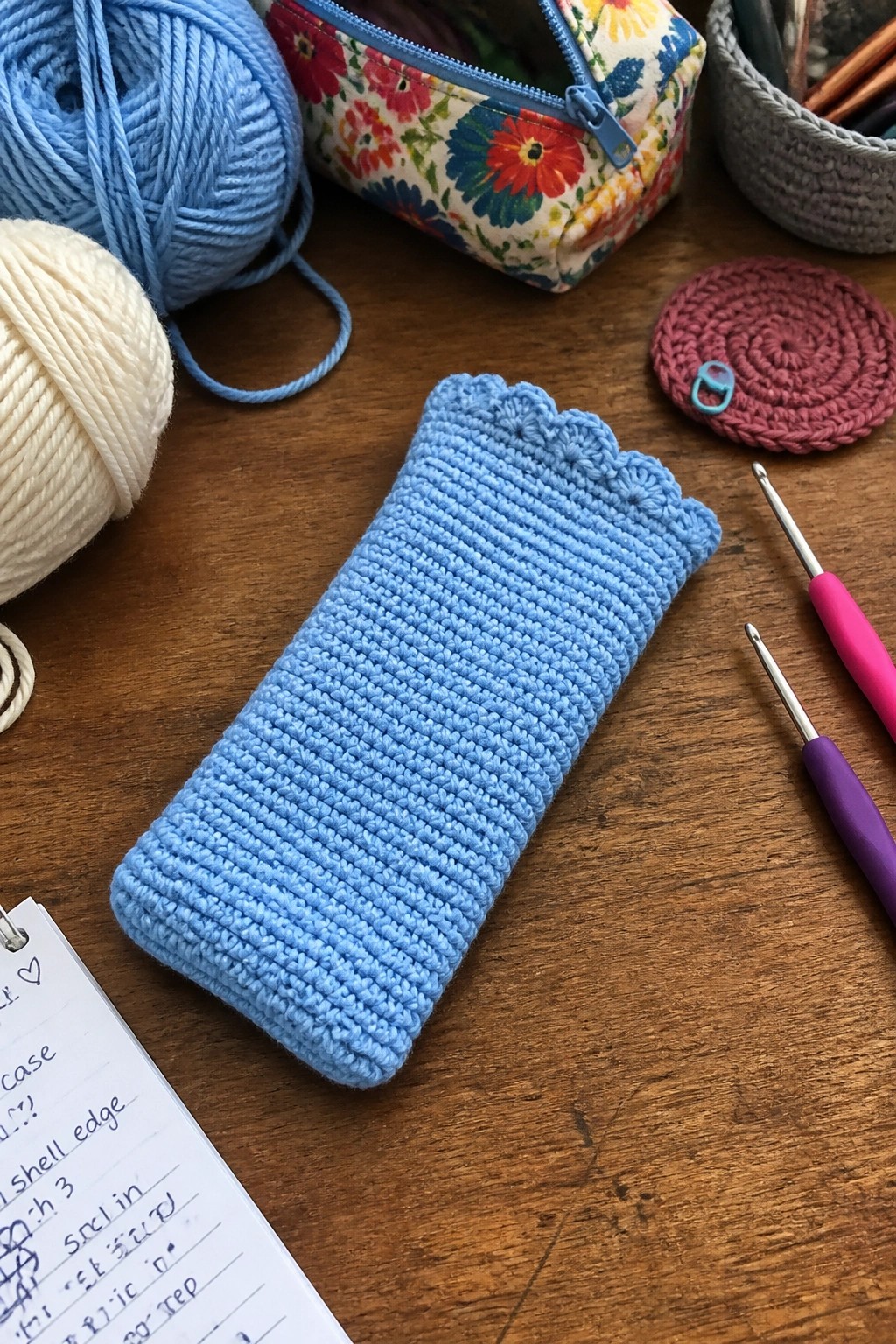 Blue crocheted case with scalloped edge on wooden table amid yarn and hooks