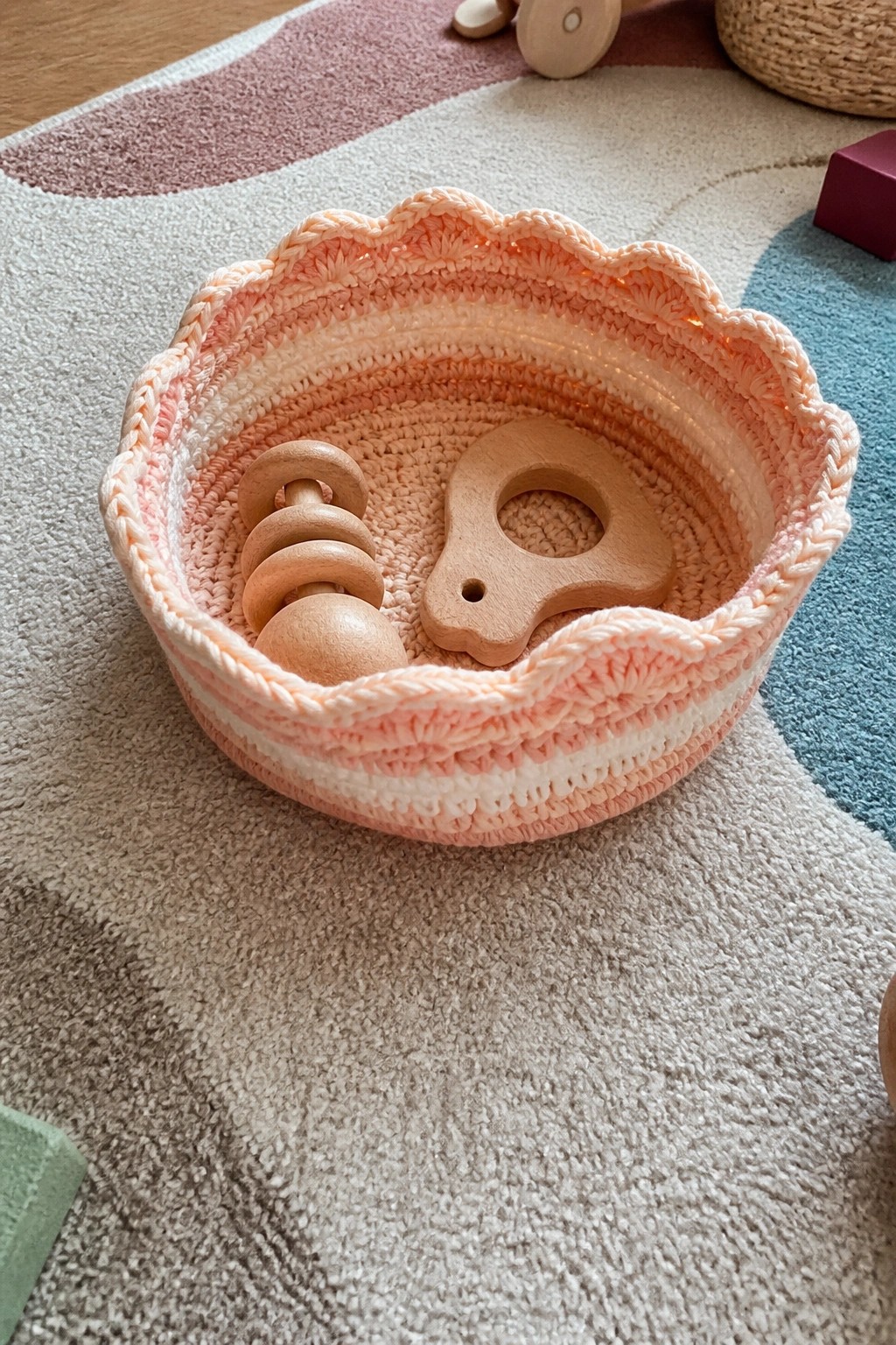 Scalloped pink crocheted basket holding wooden baby toys on textured rug.