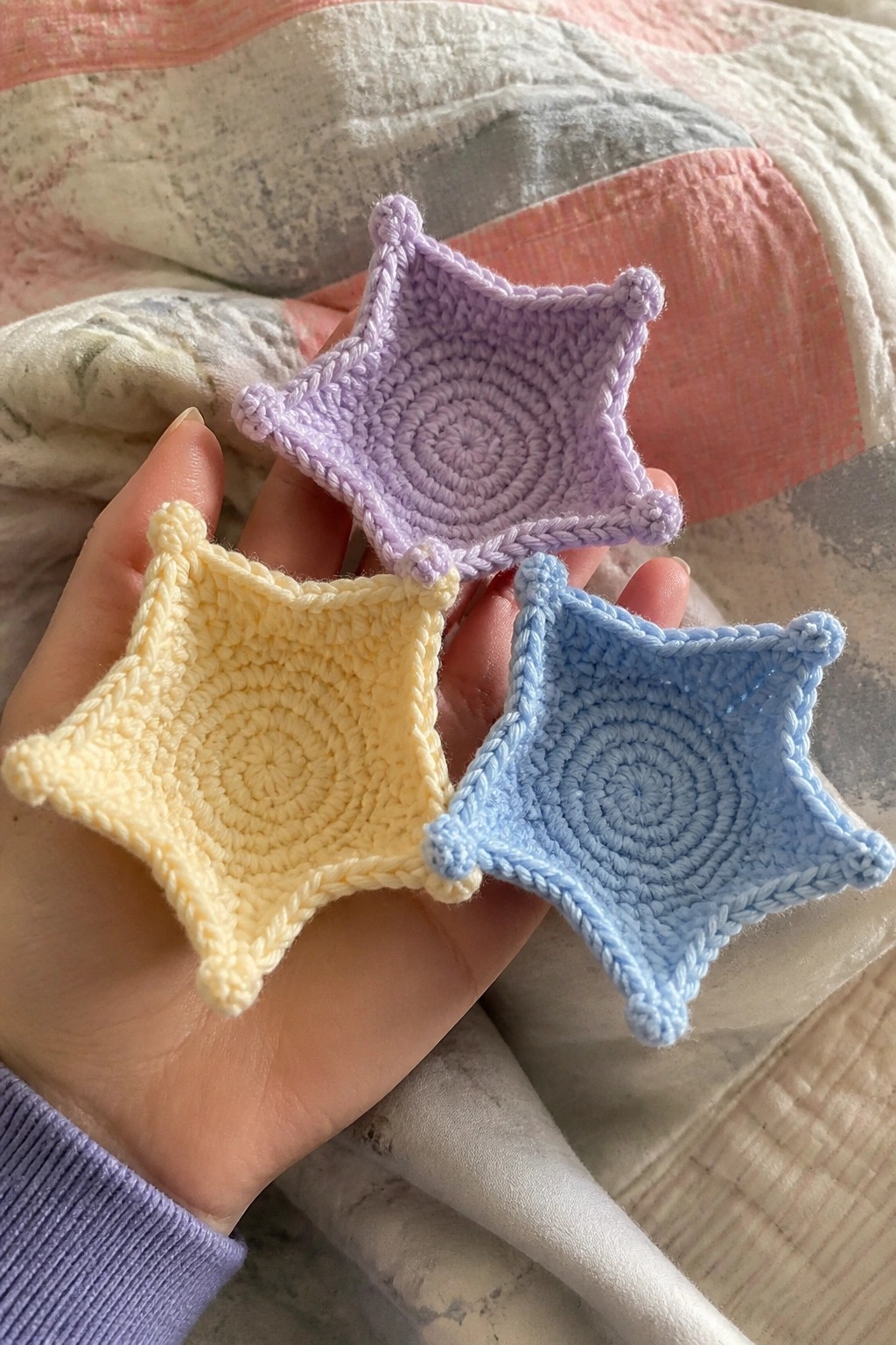 Three small crocheted star-shaped pieces in yellow, lavender, and light blue held in a hand.
