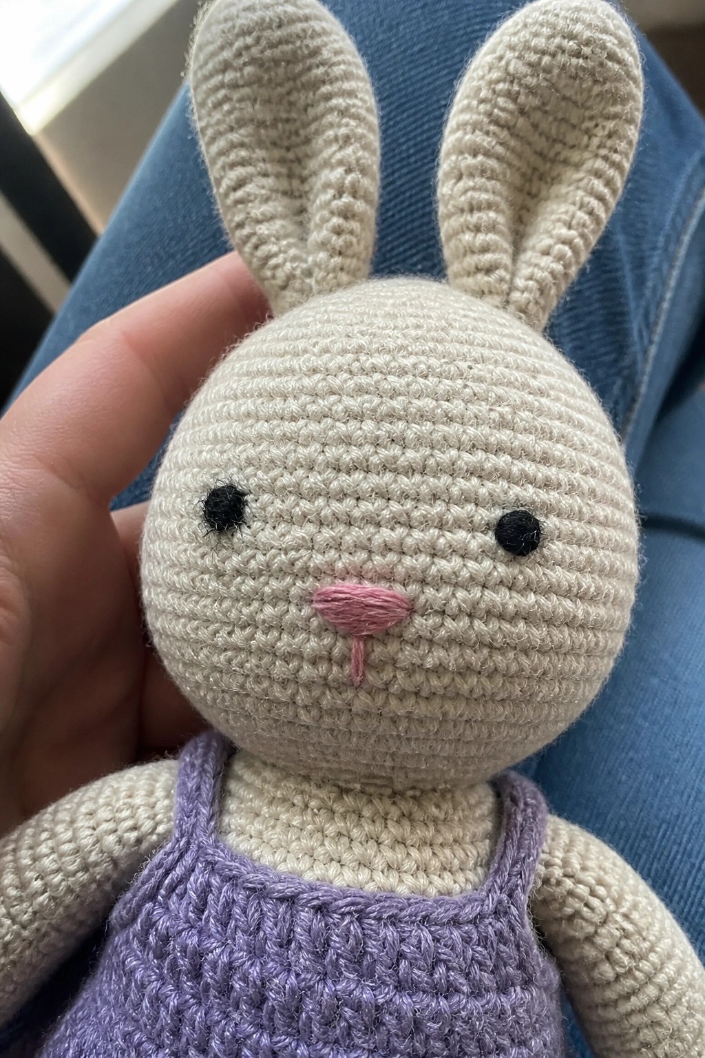 A small crocheted beige bunny amigurumi with black eyes, pink nose, and purple overalls held in a hand.