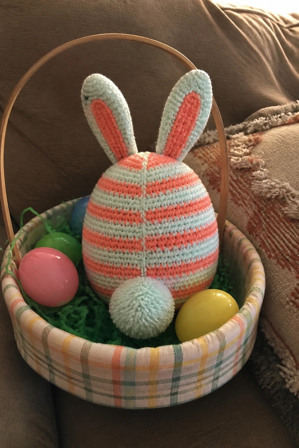 Striped crocheted bunny with pom-pom tail in plaid basket with plastic eggs.