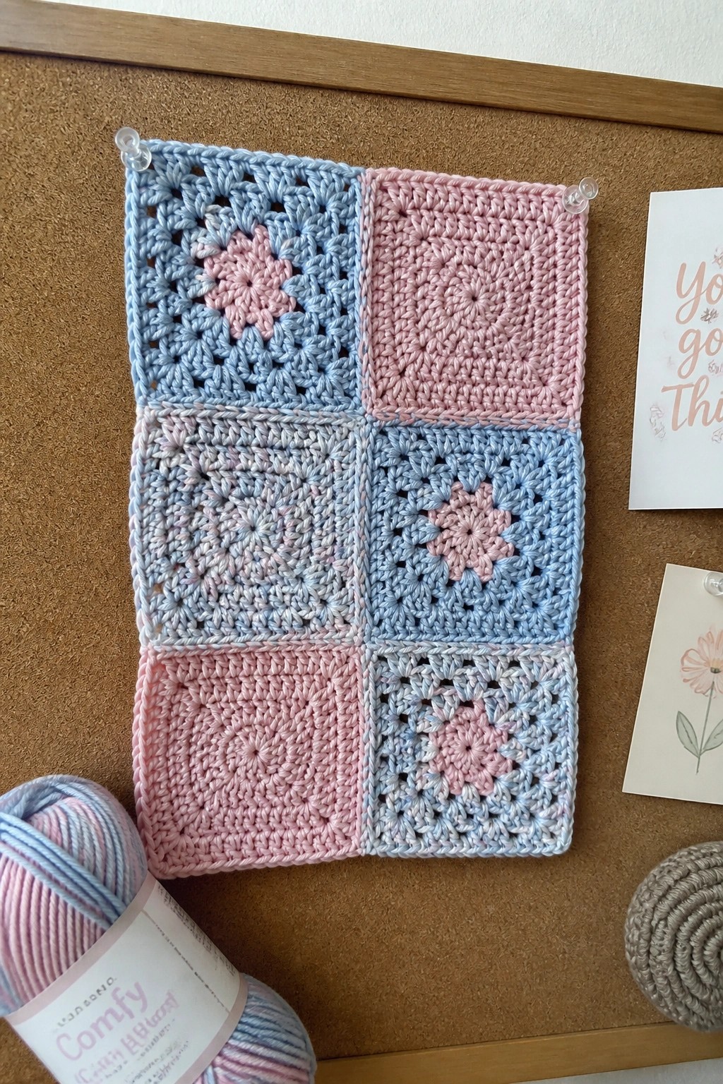 Pink and blue granny squares pinned to corkboard beside pastel yarn and cards.