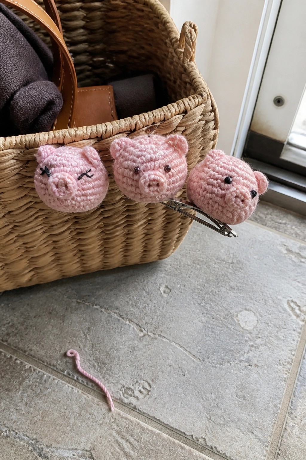 Three pink crocheted pig barrettes clipped to a woven straw basket on tiled floor.