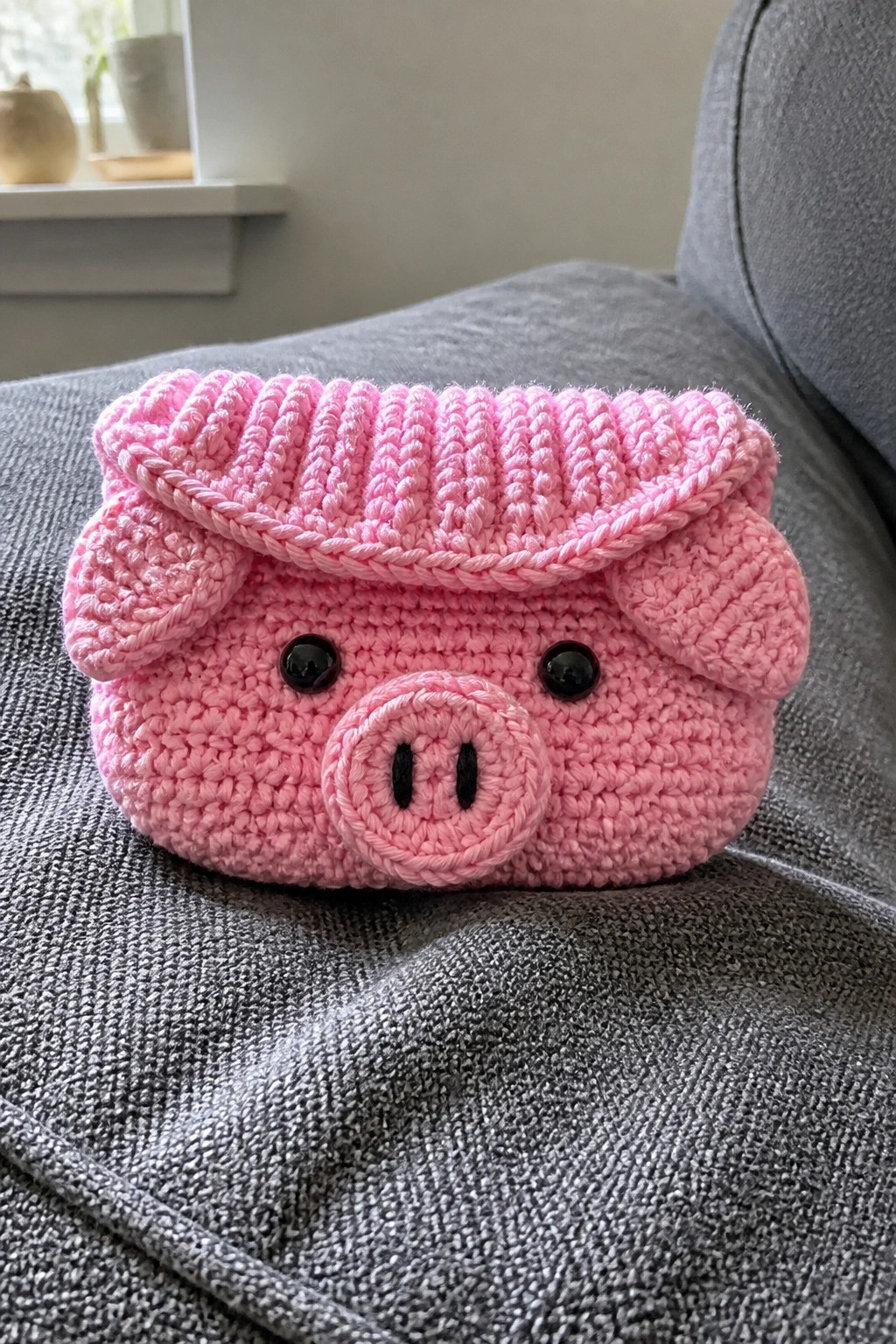 Crocheted pink pig purse with black eyes, snout, and flap on gray fabric