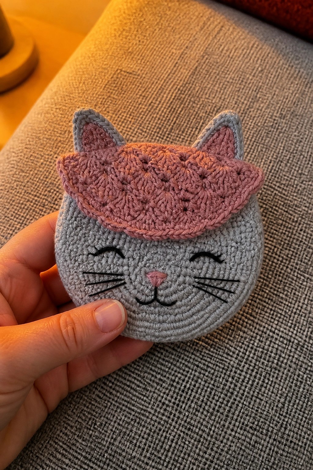 Hand holding gray crocheted cat pouch with pink hat and embroidered face