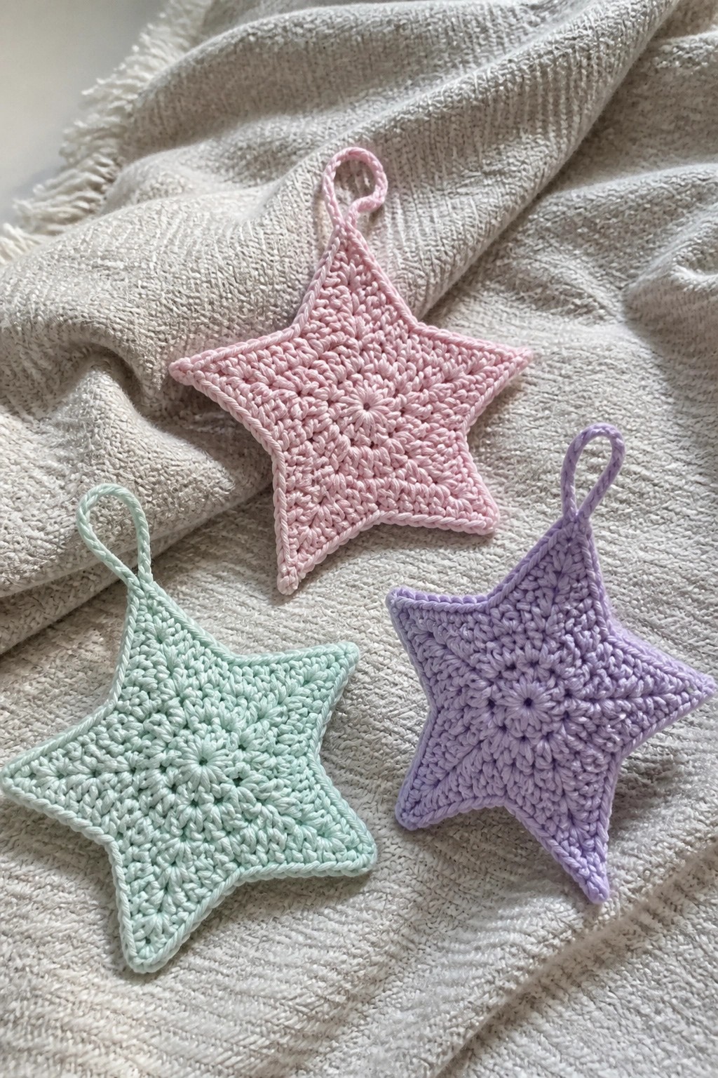 Three pastel crocheted star ornaments in pink, mint, and purple on white fabric.