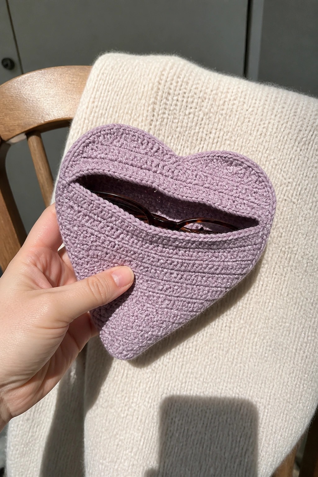 Hand holding lilac crocheted heart pouch with sunglasses on white knit sweater.