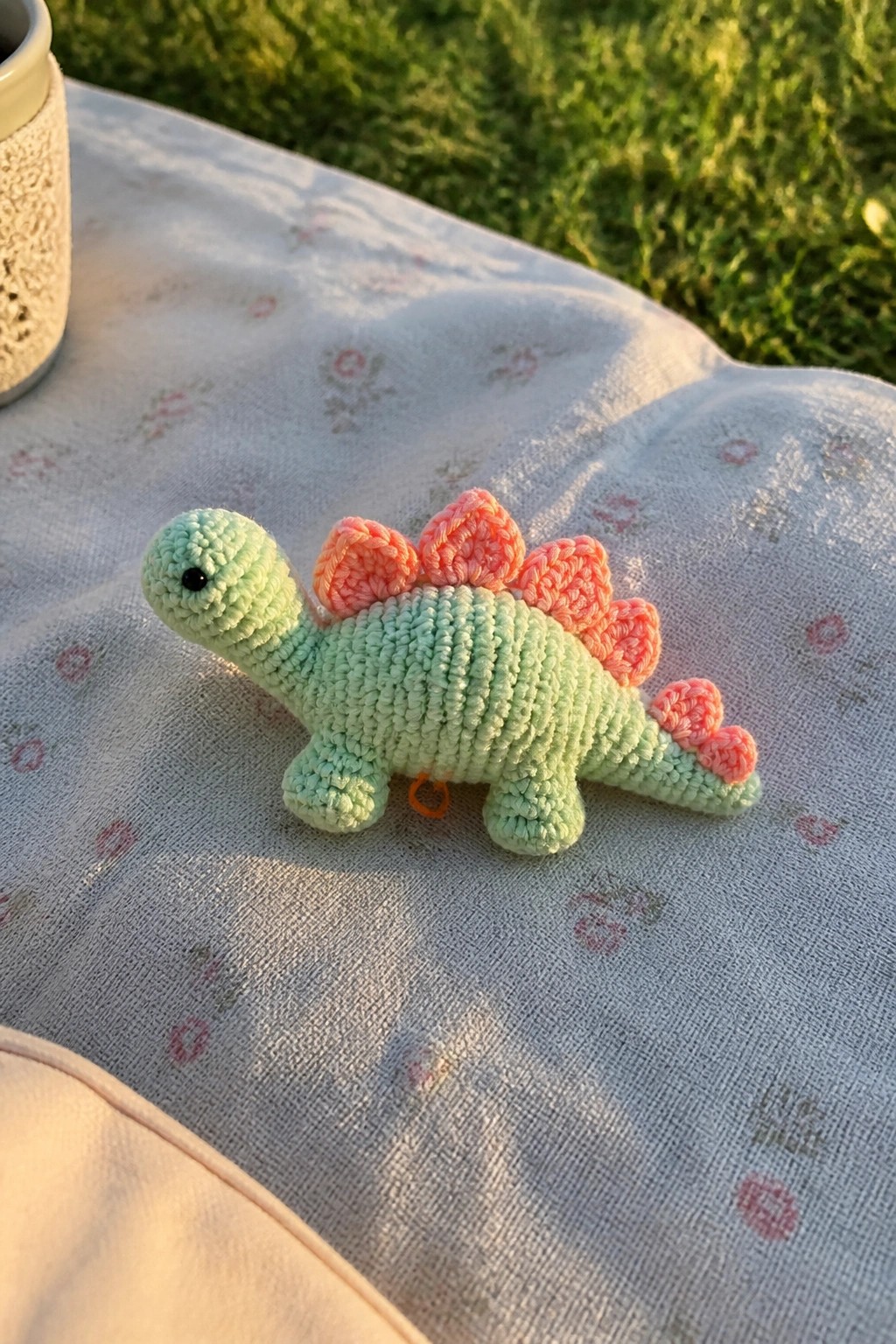 Mint green crocheted stegosaurus with pink spikes on floral fabric outdoors.