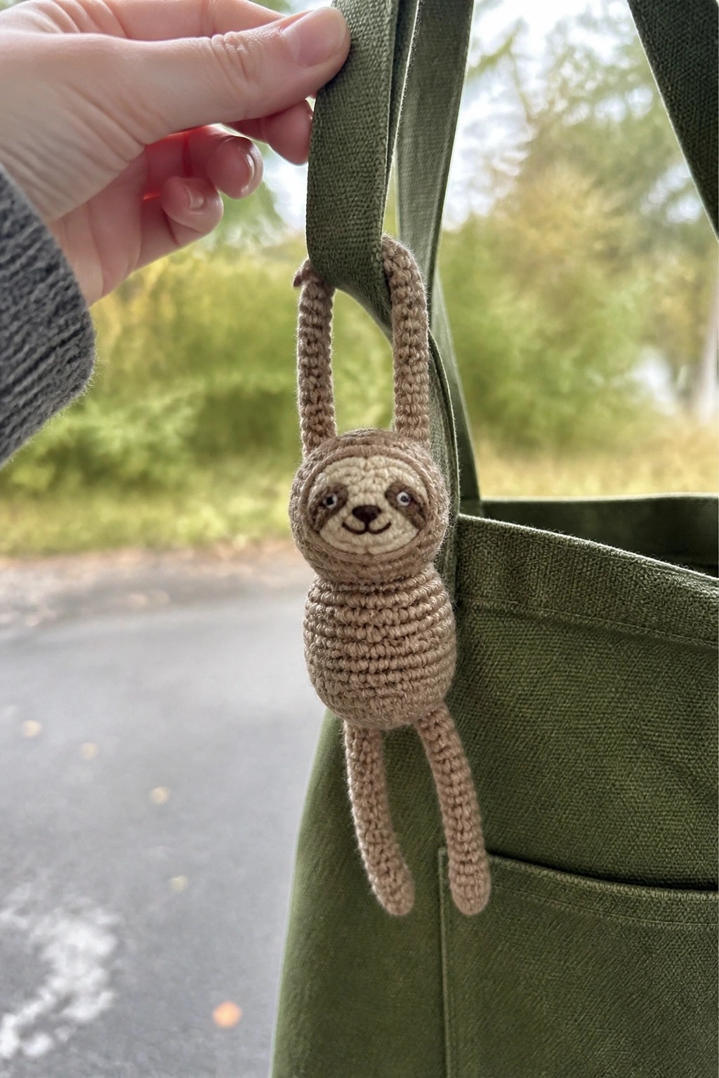 Gray-sleeved hand holds green strap with beige crocheted sloth dangling from bag.